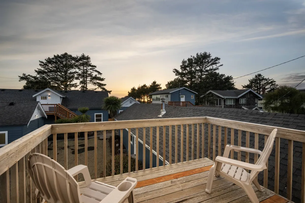 A tranquil view from a deck featuring two chairs positioned for sunset gazing. The scene captures vibrant sunset colors over the ocean, with some surrounding houses and trees visible, evoking a relaxed, romantic seaside ambiance.