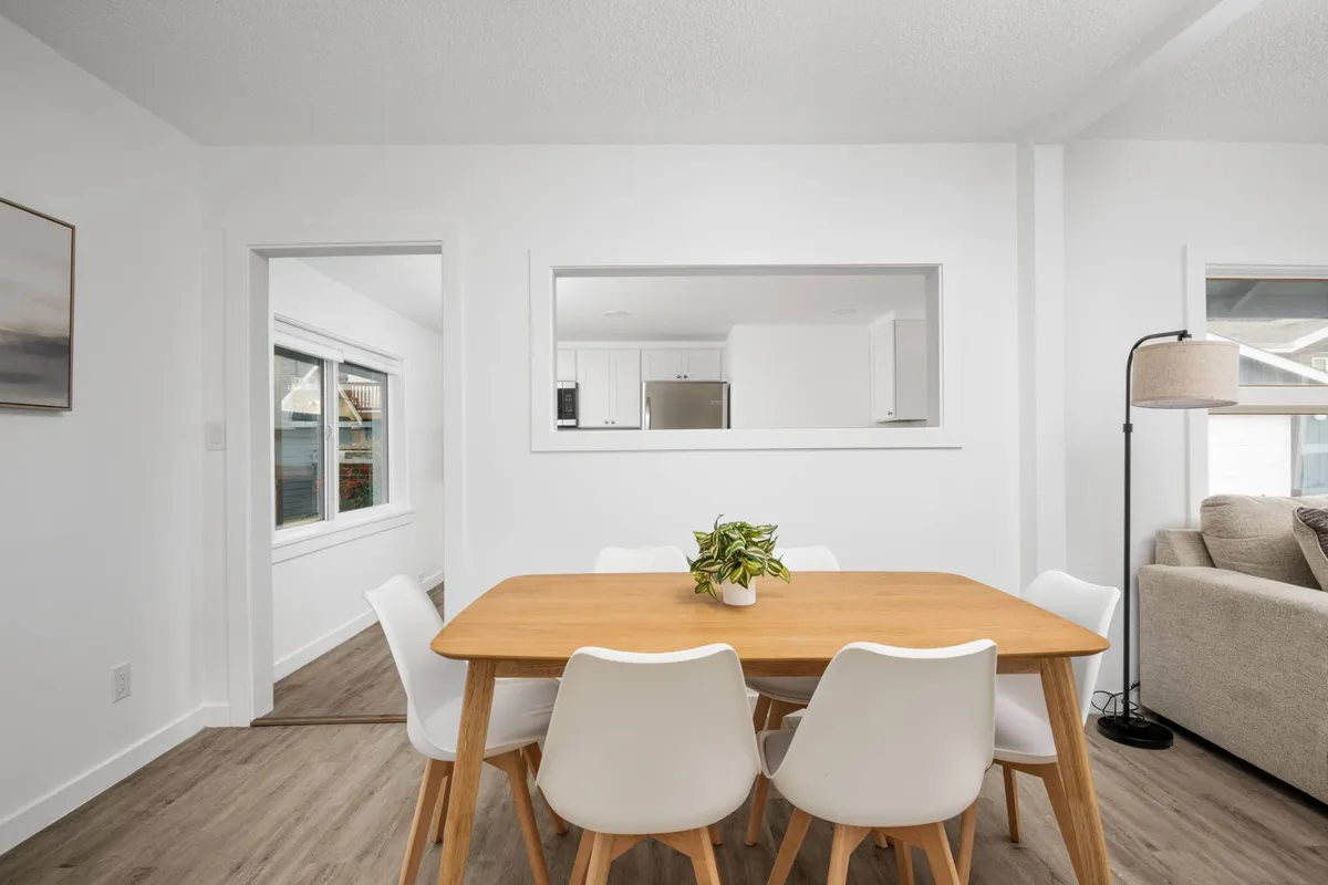 A cheerful interior with a central dining table surrounded by white chairs, an open view into the kitchen, and a soft lamp in the corner. The space is bright, modern, and inviting, ideal for group meals or casual hangouts.