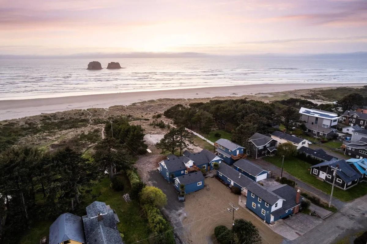 The hero image showcases the Ocean Rogue Inn condo with sweeping views of Rockaway Beach's sandy shoreline, lush greenery, and the dramatic Twin Rocks formations rising from the Pacific. The inviting exterior hints at direct beach access and a tranquil community space, with ample opportunities for seaside relaxation and adventure.