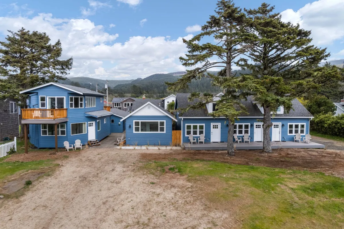 The exterior of a blue building with a deck, outdoor seating, large trees, and multiple entrances, inviting groups and families to gather, relax, or barbecue. The outdoor communal space encourages guests and their pets to unwind together after a day exploring the coastline.