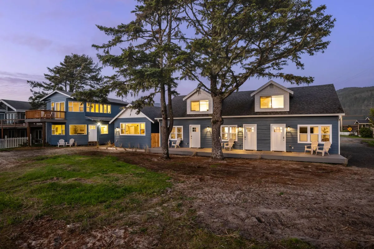 The main house and adjacent building warmly lit at twilight, framed by tall trees. The inviting scene features porches and balconies ideal for relaxing or gathering as a group under the stars.