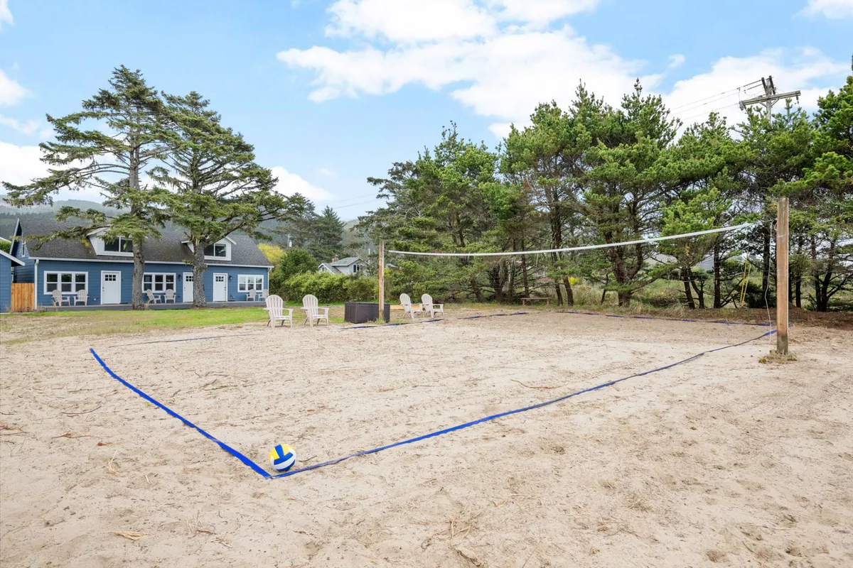 The volleyball court is set on sand, featuring a net, adirondack chairs for spectators or relaxation, and the blue houses in the background. Groups are seen playing volleyball and socializing, creating a lively and playful environment.
