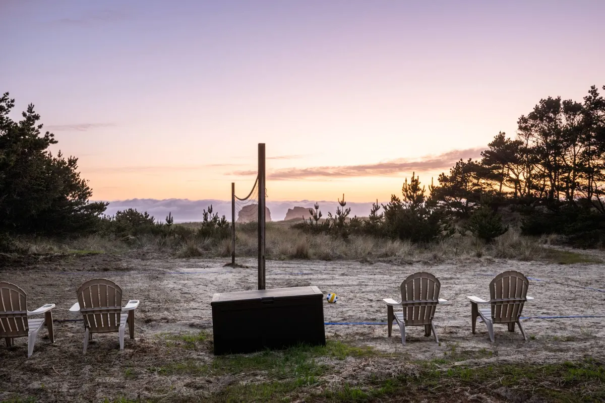 Wooden chairs are set up beside a volleyball net, looking out toward a sunset horizon with distant rock formations. The scene is tranquil yet social, suggesting both active play and peaceful moments watching the sunset together.