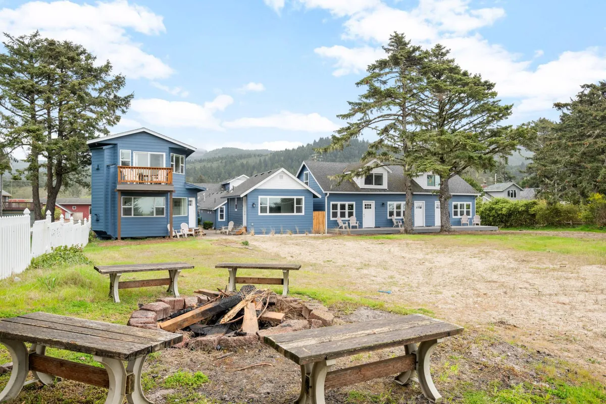 This image captures the fire pit surrounded by green grass and blue houses, with a large tree providing shade. Several people are gathered around the fire pit, enjoying a communal and welcoming atmosphere that invites shared stories and relaxation.