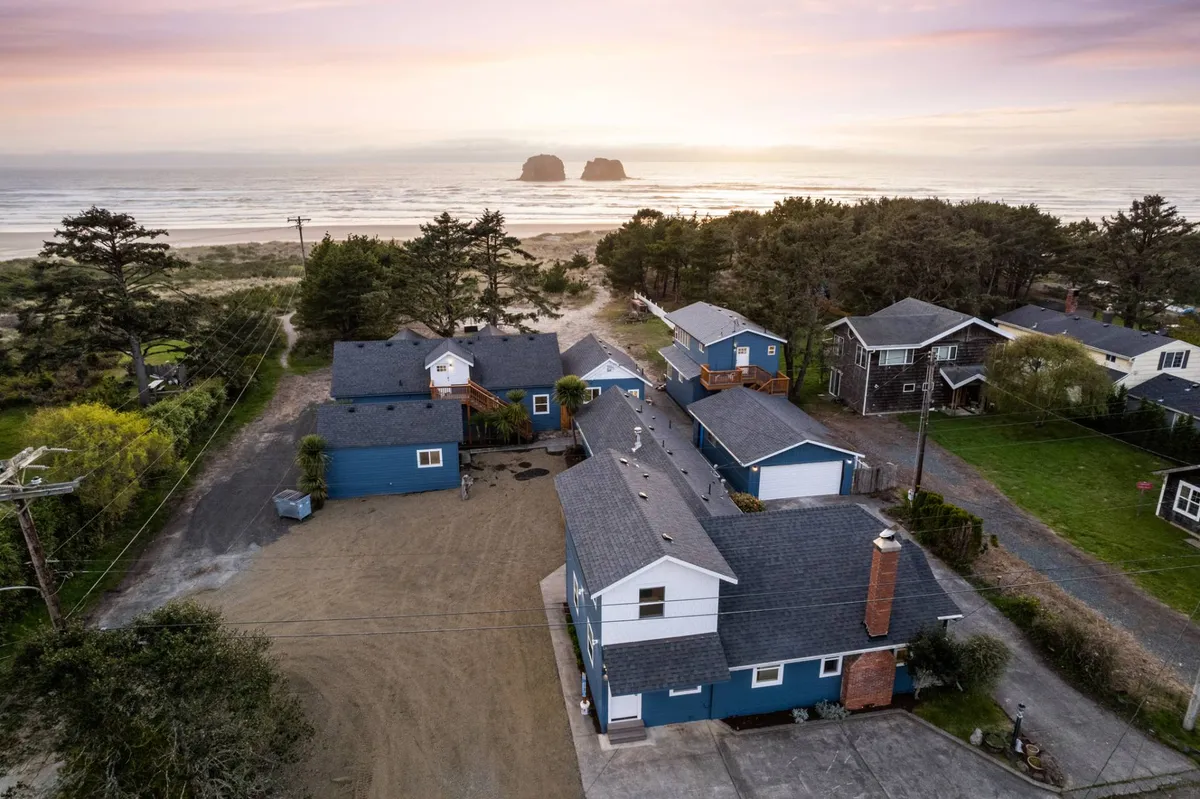 A wide view of Rockaway Beach’s sandy shoreline, with calm ocean waves, distant rock formations (possibly the iconic Twin Rocks Arch), and a few houses and trees along the edge of the beach. The open space is perfect for families to enjoy beachcombing, sandcastle building, and sunset watching.
